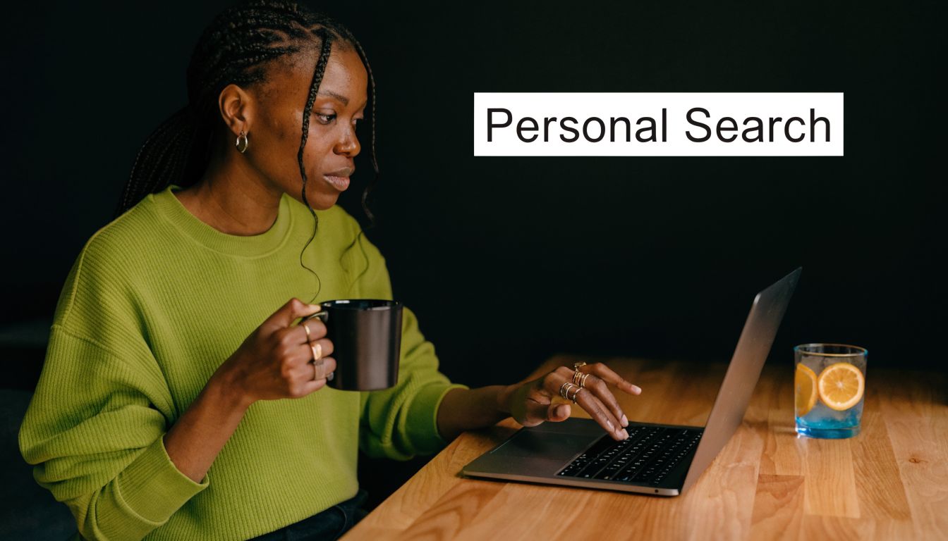 A woman in a green sweater uses a laptop while holding a mug at a wooden table.